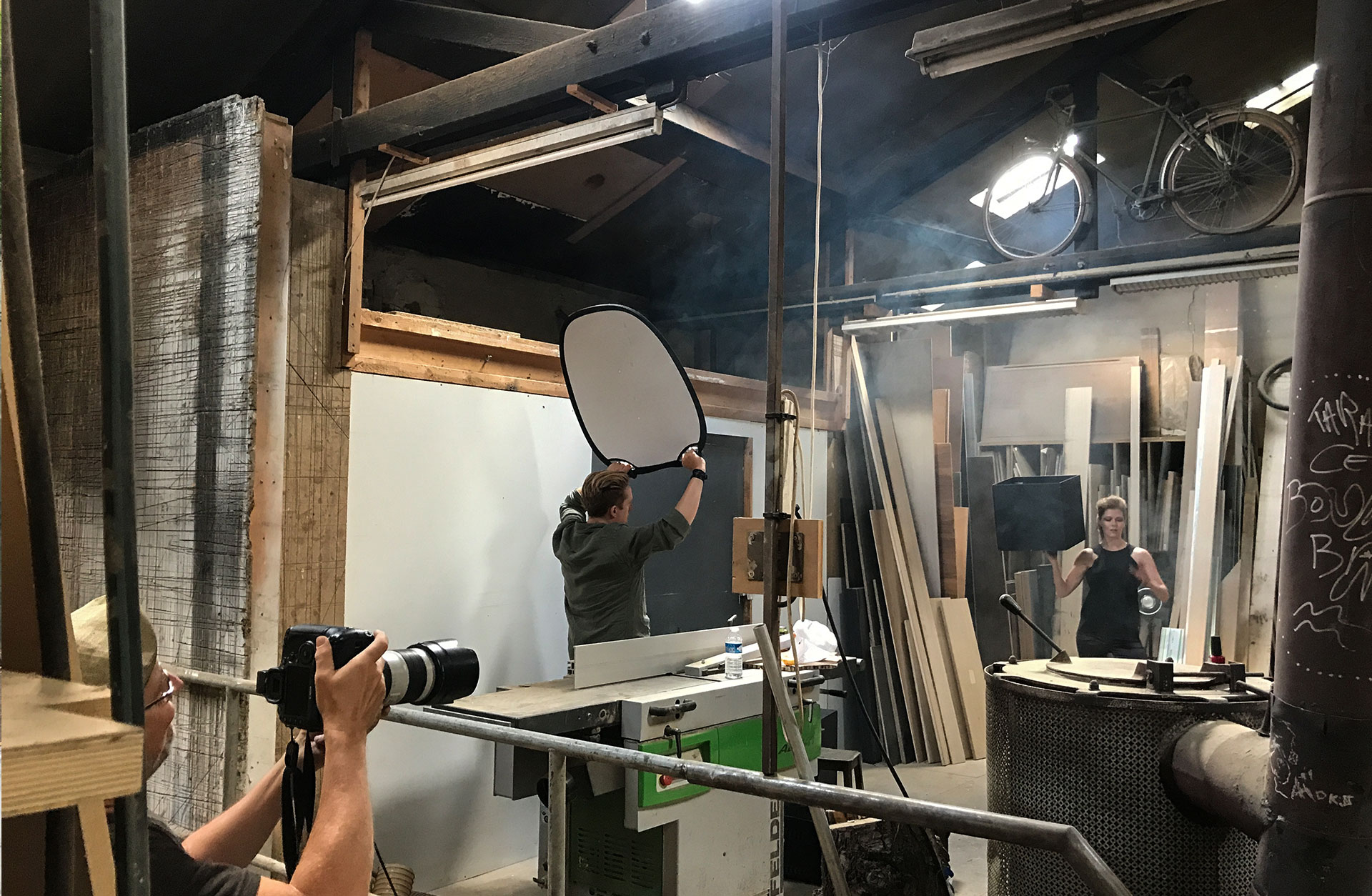 Mats Bäcker fotograferar Barbara som poserar med den svarta lådan i en hand. Linus håller en reflektor. miljön är stökig.