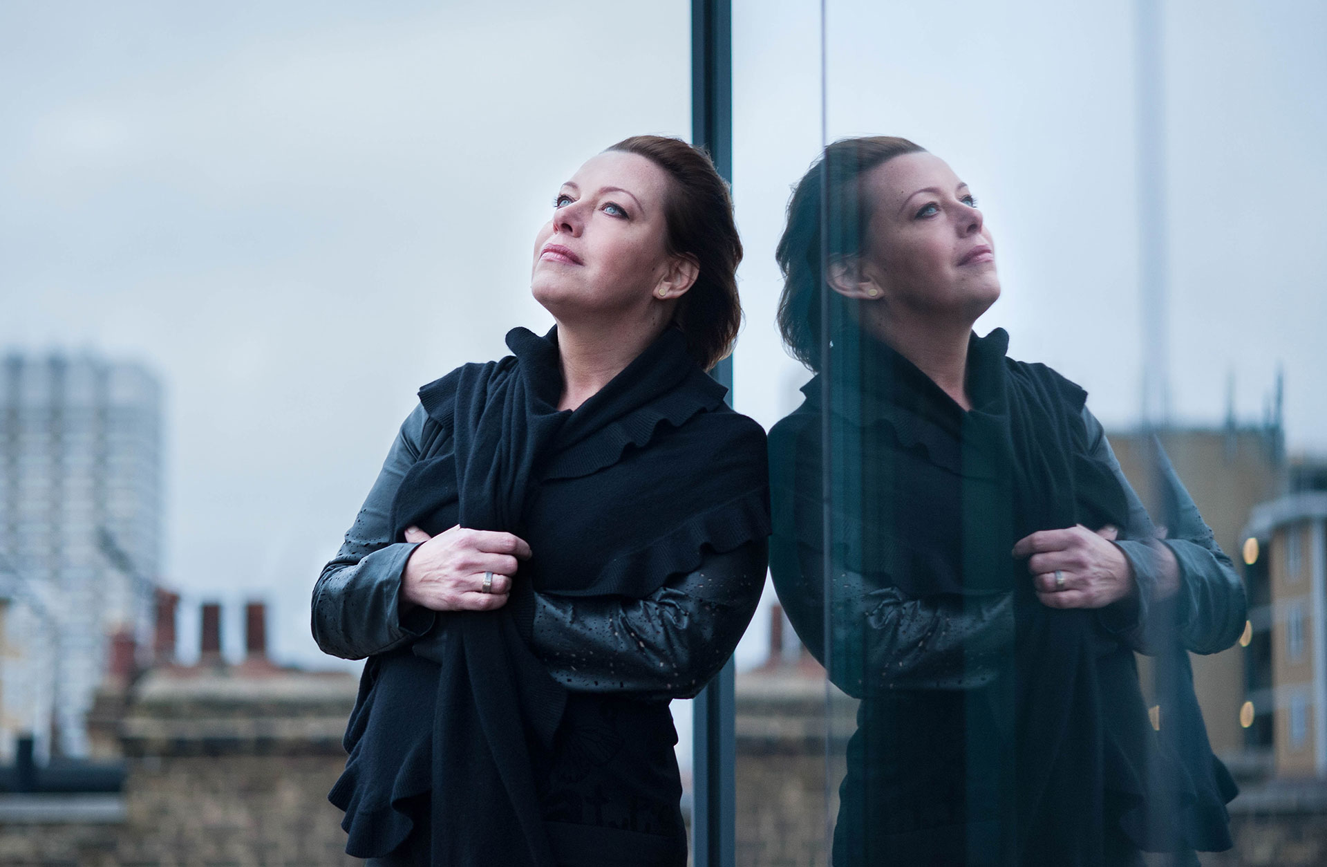 Nina Stemme står lutad mot ett fönster, utomhus. Hennes spegelbild reflekteras i fönstret. 