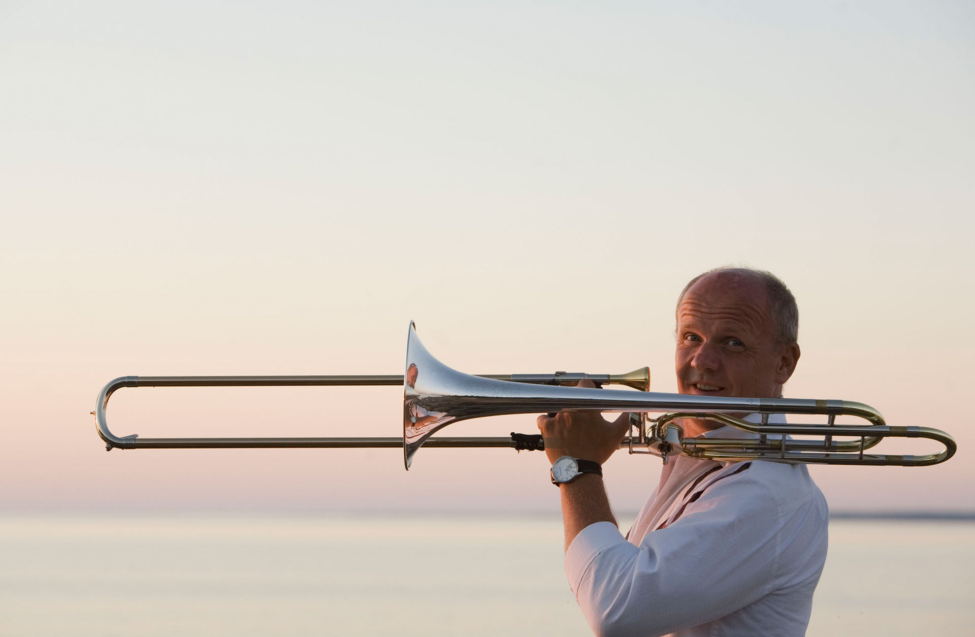 Christian Lindberg håller upp trombonen och tittar in i kameran. Havet syns i bakgrunden.