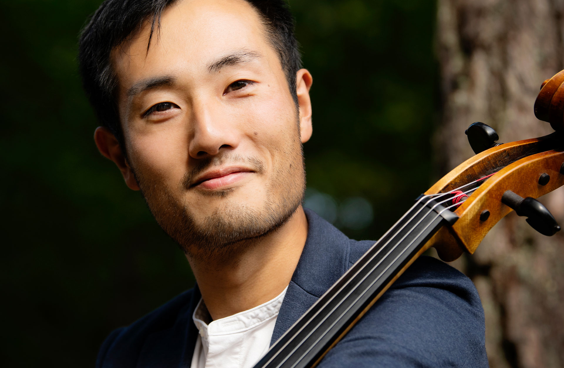 Jun Sasaki sitter utomhus mot ett träd med sin cello och tittar in i kameran.