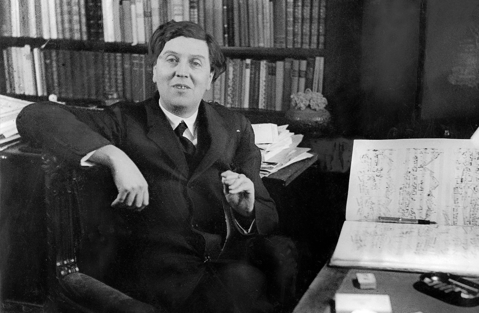 Alban Berg med cigarr i handen vid sitt skrivbord. Noter ligger på bordet och han tittar in i kameran.