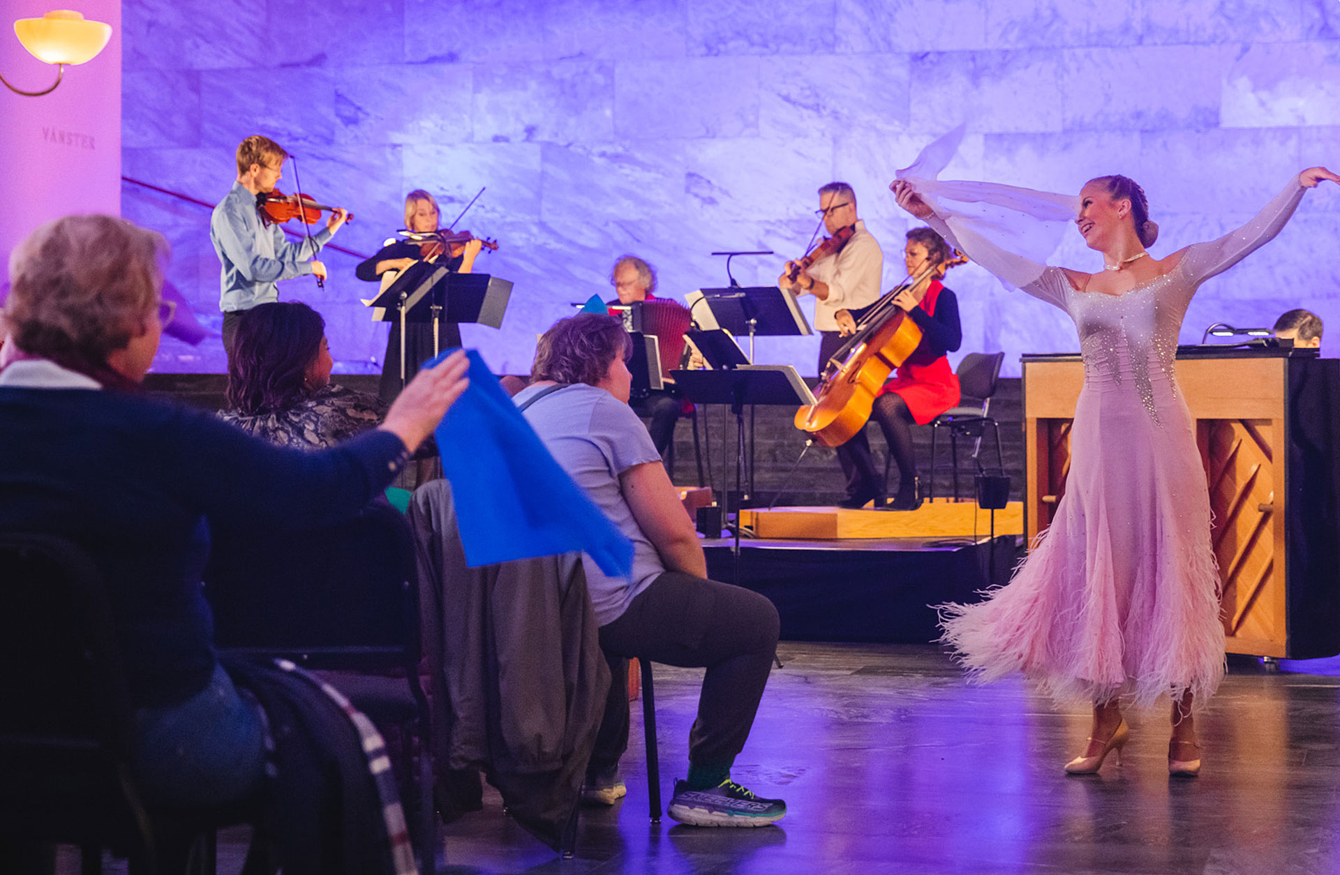 En dansare i vacker klänning dansar vid sidan av publiken med en sjal i handen, på scenen syns fem musiker.