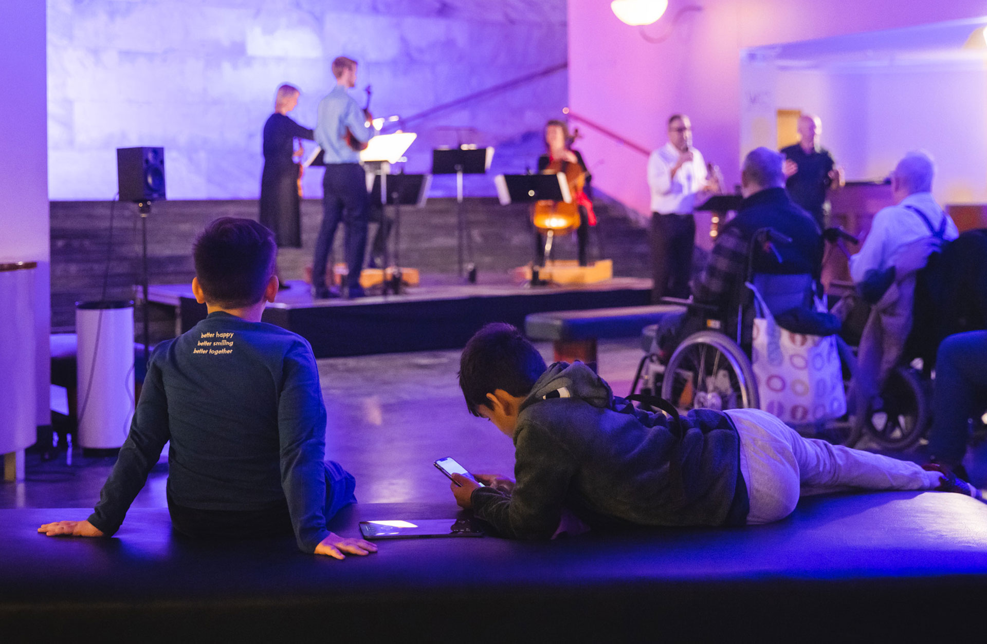 Två barn i publiken ligger och vilar på en mjuk bänk medan de lyssnar på musikerna på scenen.