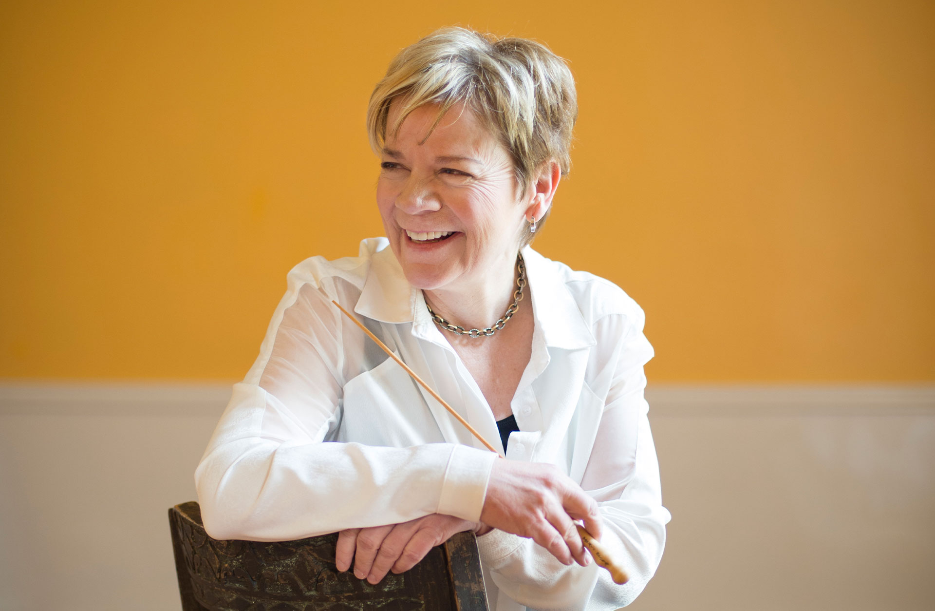 Marin Alsop i vit blus sitter mot en gul vägg och skrattar.