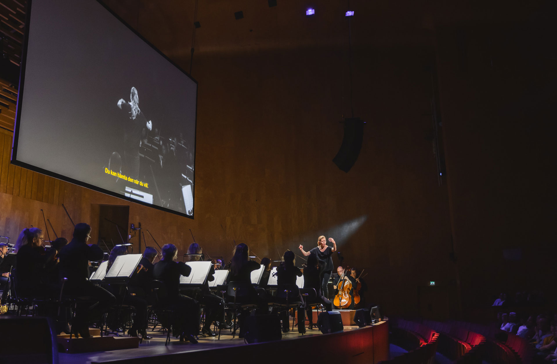 Barbara Hannigan står på pulten och dirigerar orkestern. Ovanför orkestern ser man Barbara projiceras på en skärm.