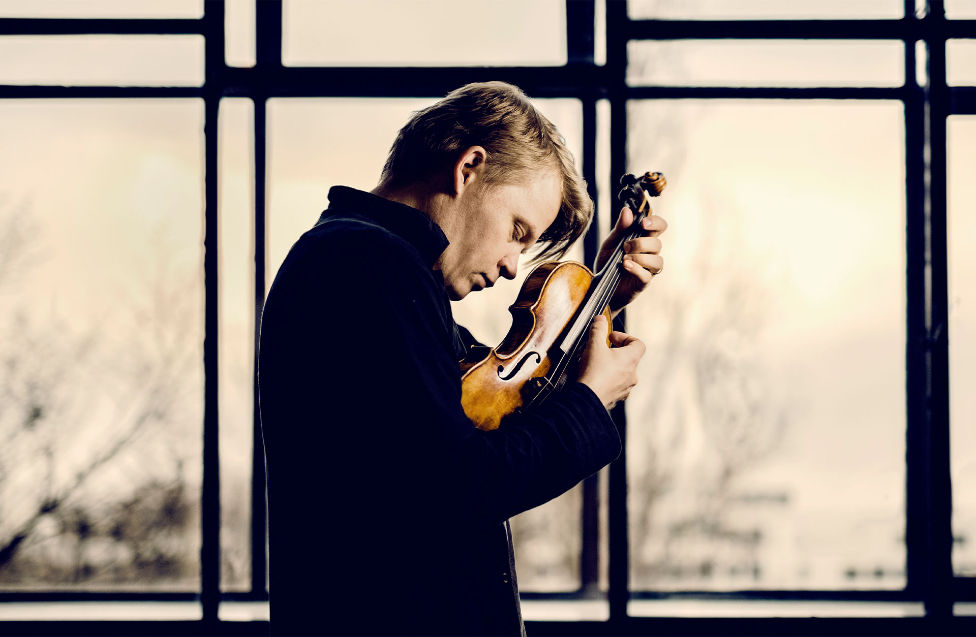 Pekka Kuusisto står i profil framför ett fönster och knäpper på strängarna på sin fiol.