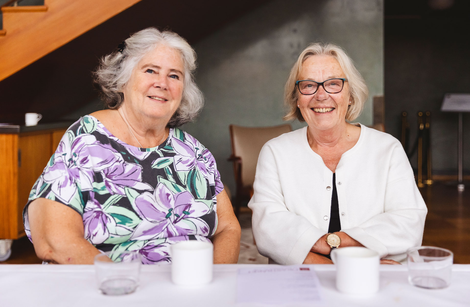 Två damer sitter vid ett fikabord. De ler och tittar in i kameran.