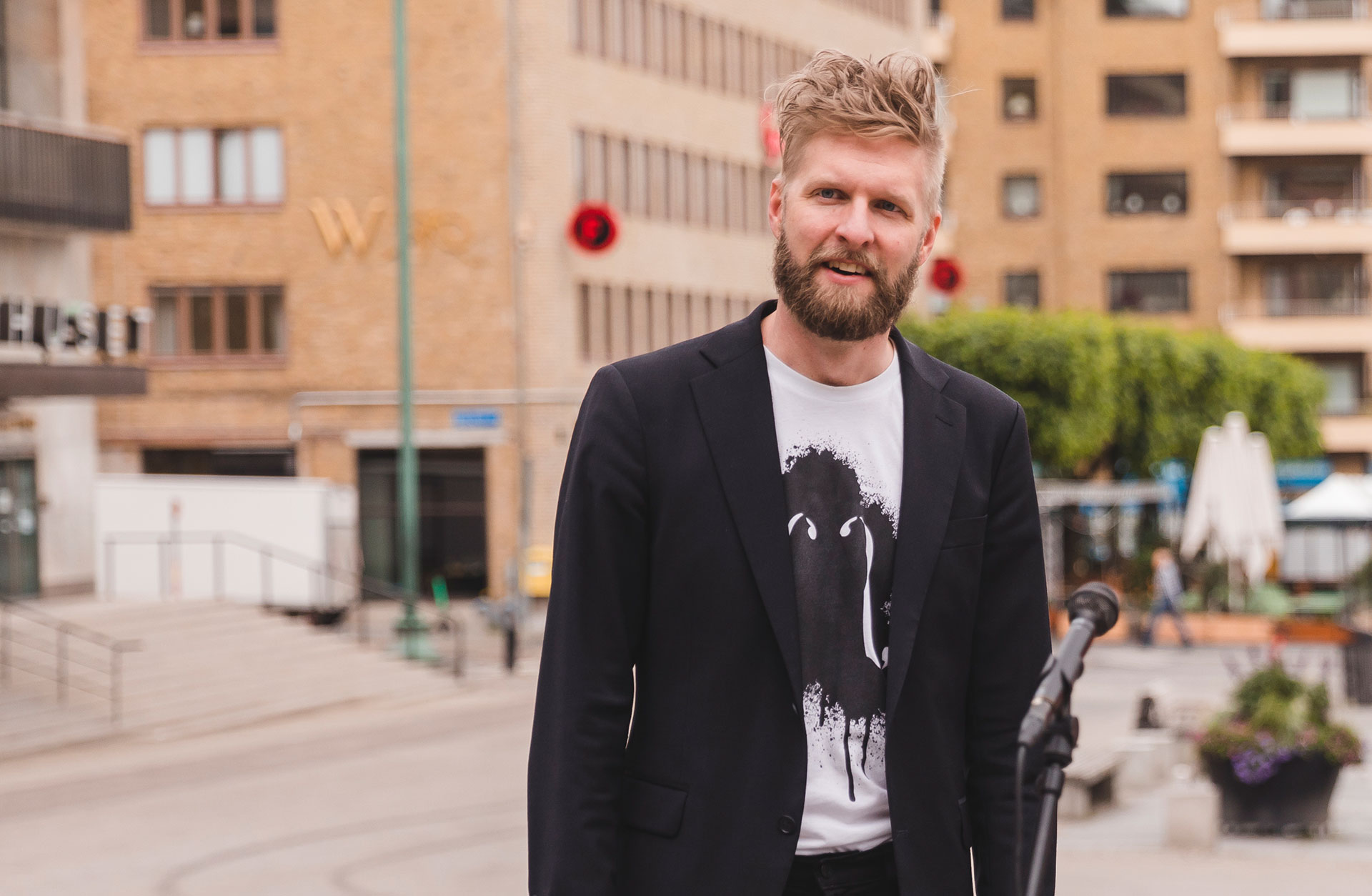 Johan Andersson Duraffourdstår på Götaplatsen i en vit t-shirt med Side by Side-logga på bröstet och en svart kavaj. Han har kortklippt blont hår med busig lugg och skägg. Han är glad.
