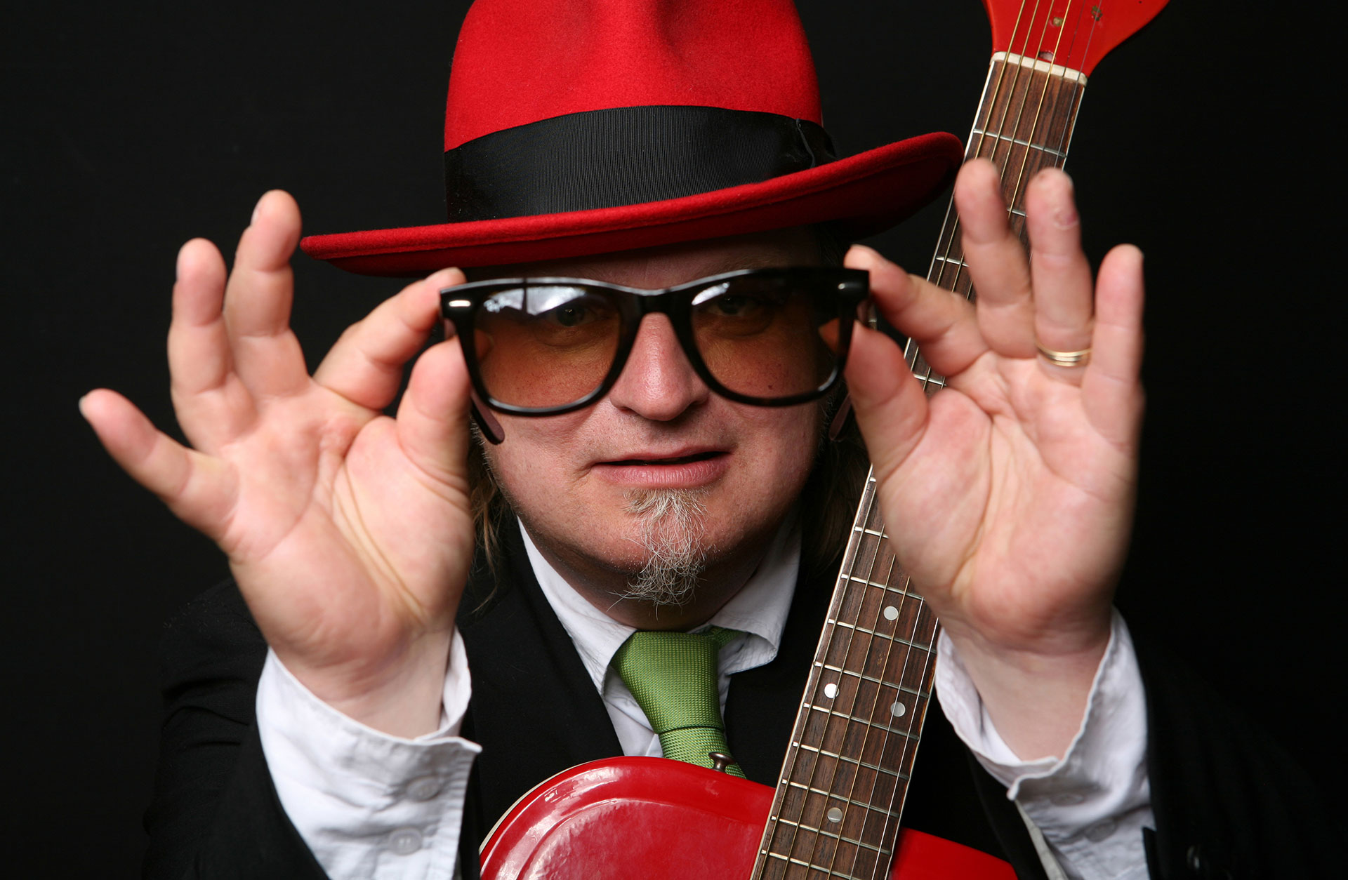 Närbild av Knut Reiersrud med sin gitarr. Han är klädd i vit skjorta, grön slips och röd hatt och håller ett par svarta solglasögon framför ansiktet.