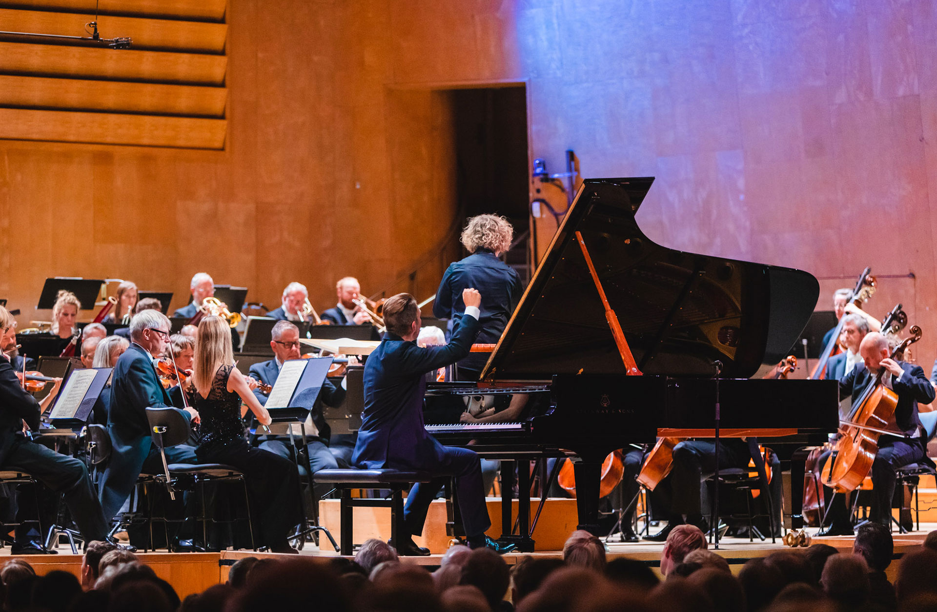Pianist i engagerad pose vid flygeln med orketser och dirigent i bakrgunden.