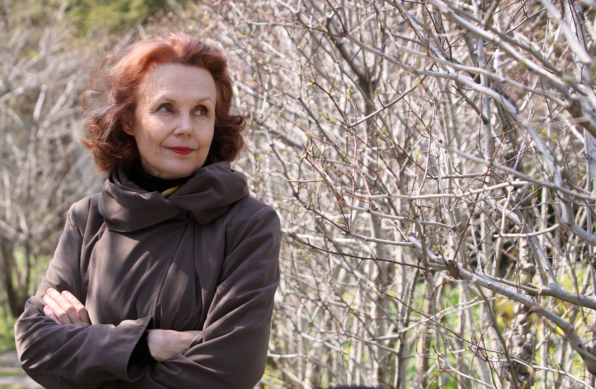 Kaija Saariaho står framför ett buskage utomhus.. Hon ser nöjd ut.