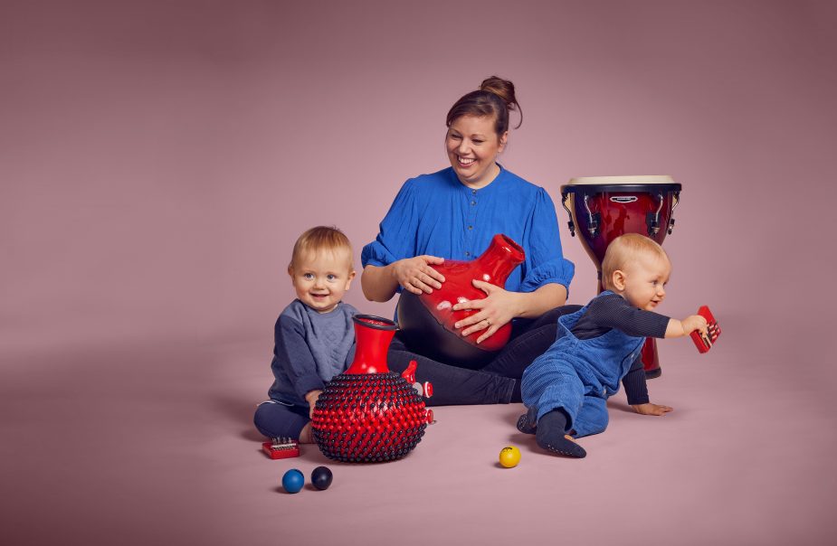 En kvinna sitter med två bebisar runt sig och de leker med instrument. De har blåa kläder mot ljusrosa bakgrund.