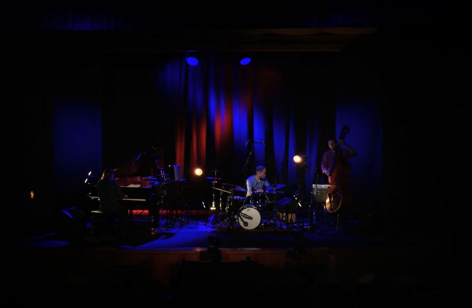 3 musiker står på scen i stenhammarsalen, det är mörkt och upplyst mitt på scen med blått ljus.