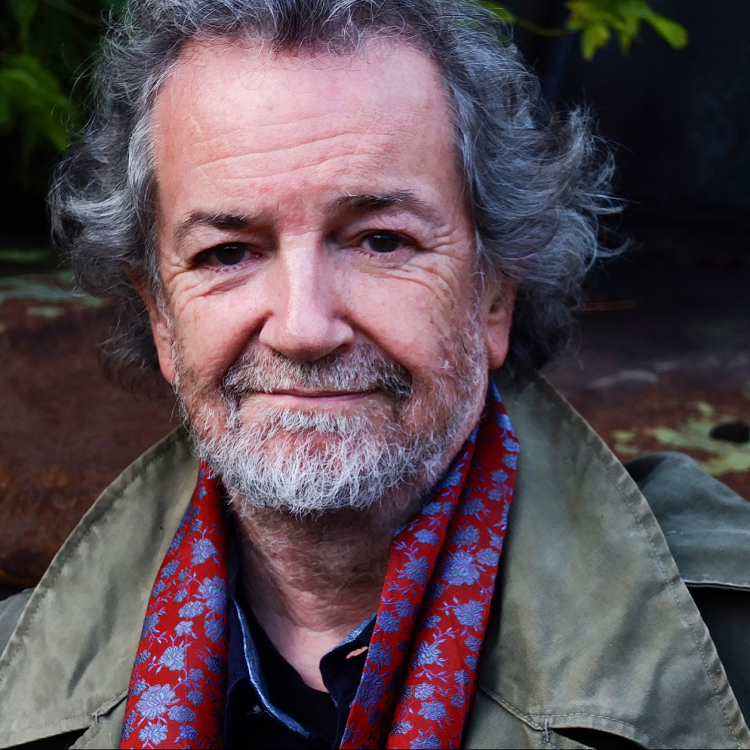 Porträtt av Andy Irvine, en äldre man med vänligt leende. Han har grått hår och skägg och ler mot kameran.