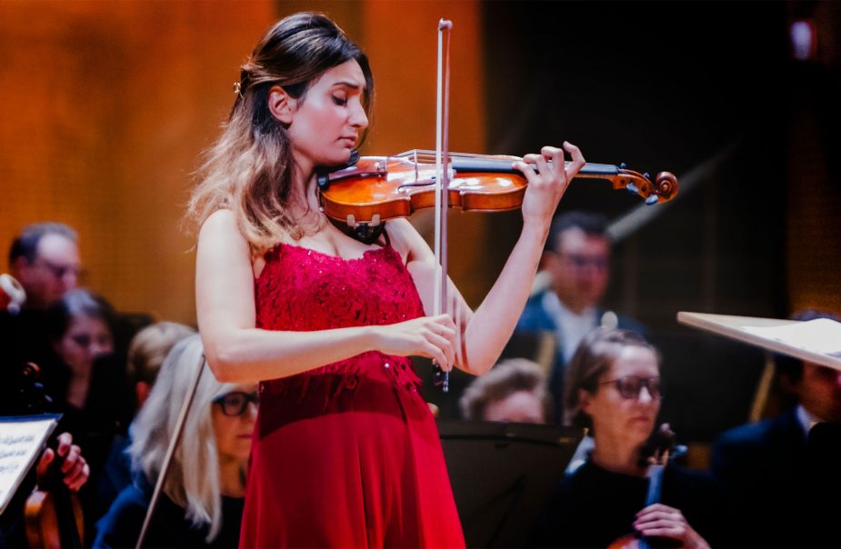En ung kvinna i röd klänning spelar violin framför en orkester.