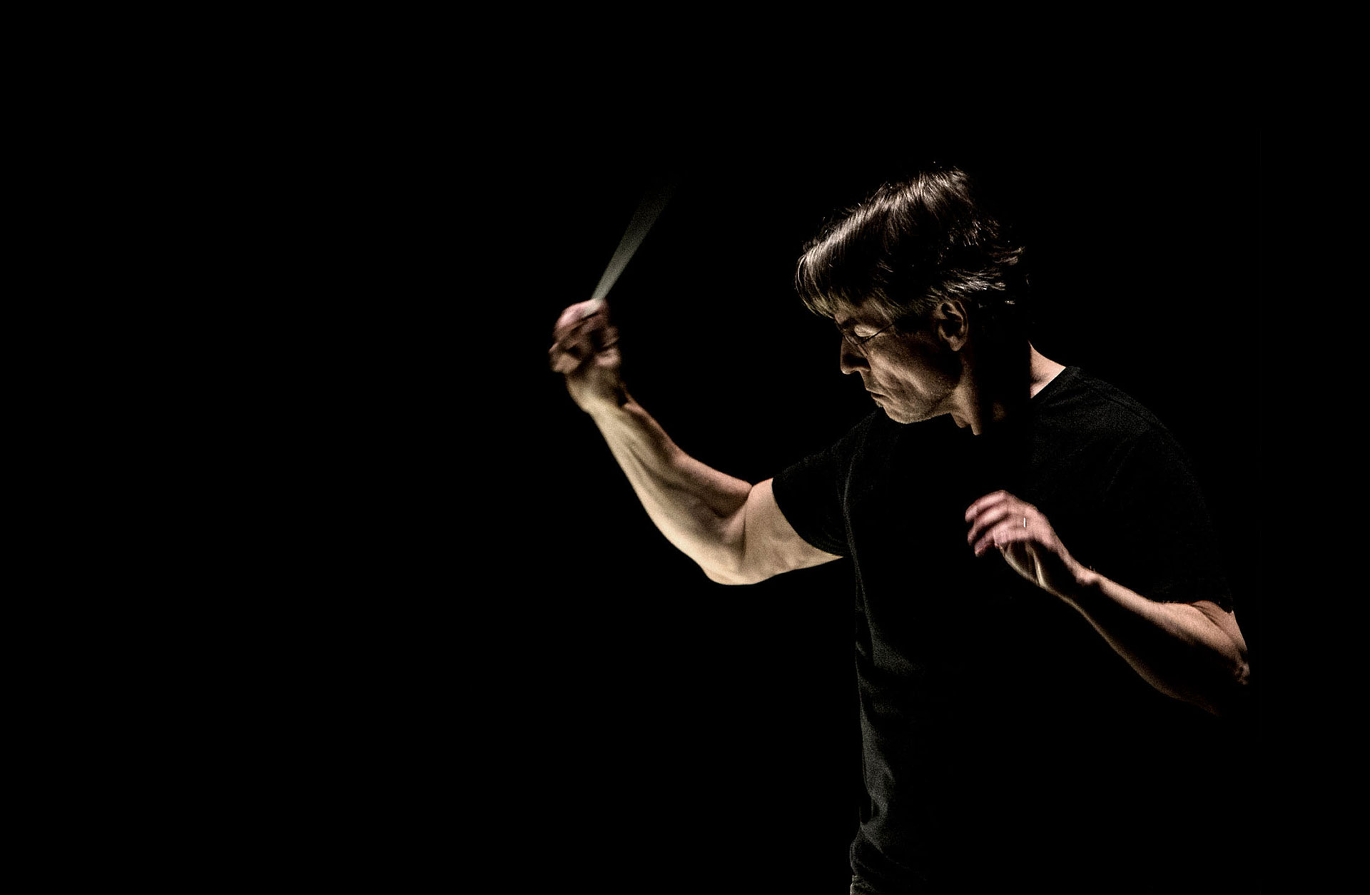Esa-Pekka Salonen, klädd i svart T-shirt, står mot en mörk bakgrund och dirigerar. Man ser honom från sidan.