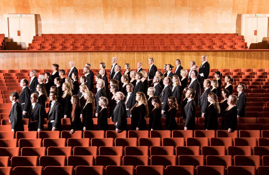 En gruppbild på många personer som står samlade i raderna mellan stolarna på parkett. De är konsertklädda och alla står uppställda i profil.