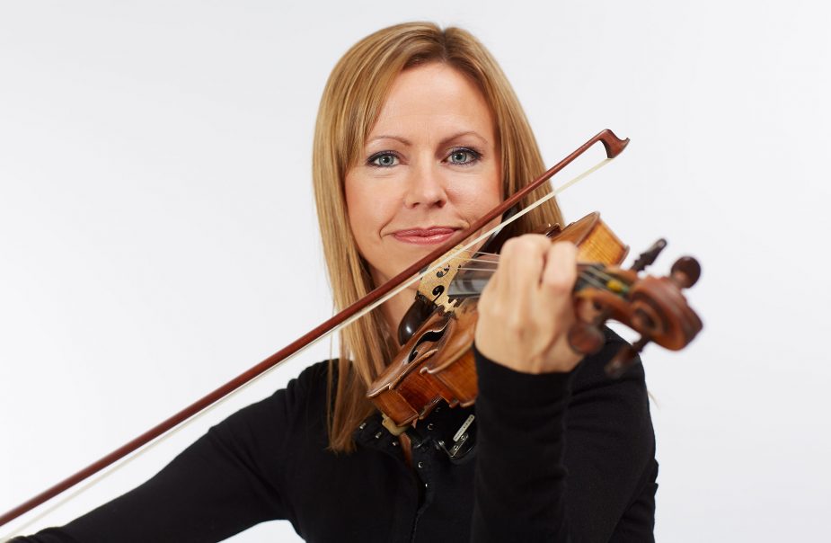 Halvkroppsporträtt av Sara Trobäck som spelar på sin violin. Hon tittar rakt in i kameran med sina blå ögon. Hon ler.