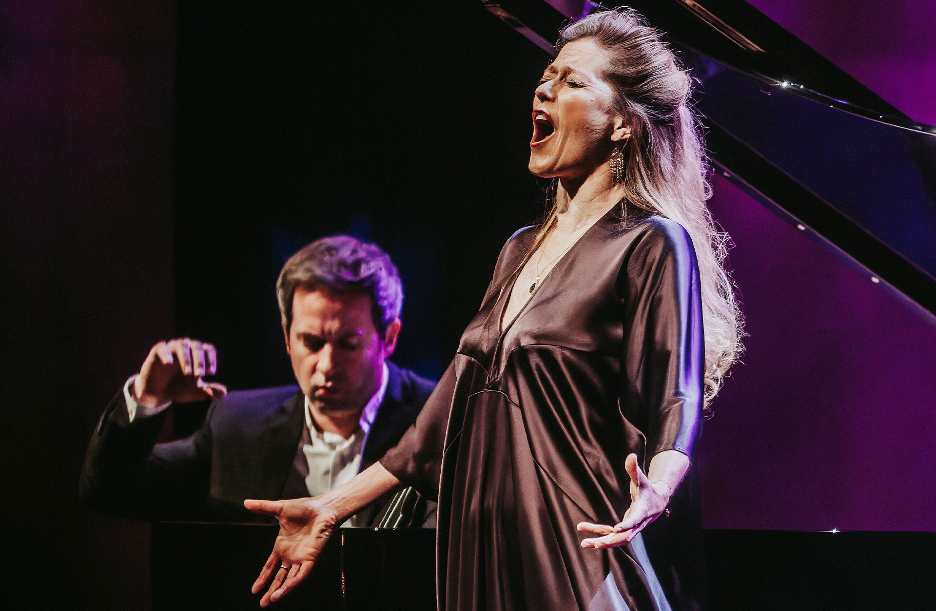 En actionbild under en konsert där En pianist spelar i bakgrunden och en kvinna i glansig klännig sjunger inlevelsefullt . Hon blundar.