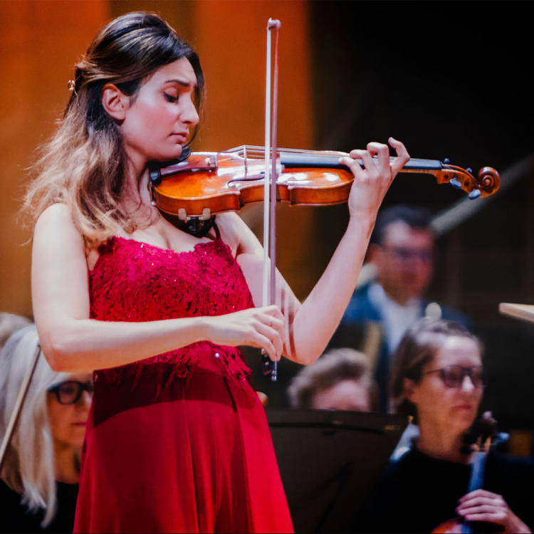 En ung kvinna i röd klänning spelar violin framför en orkester.