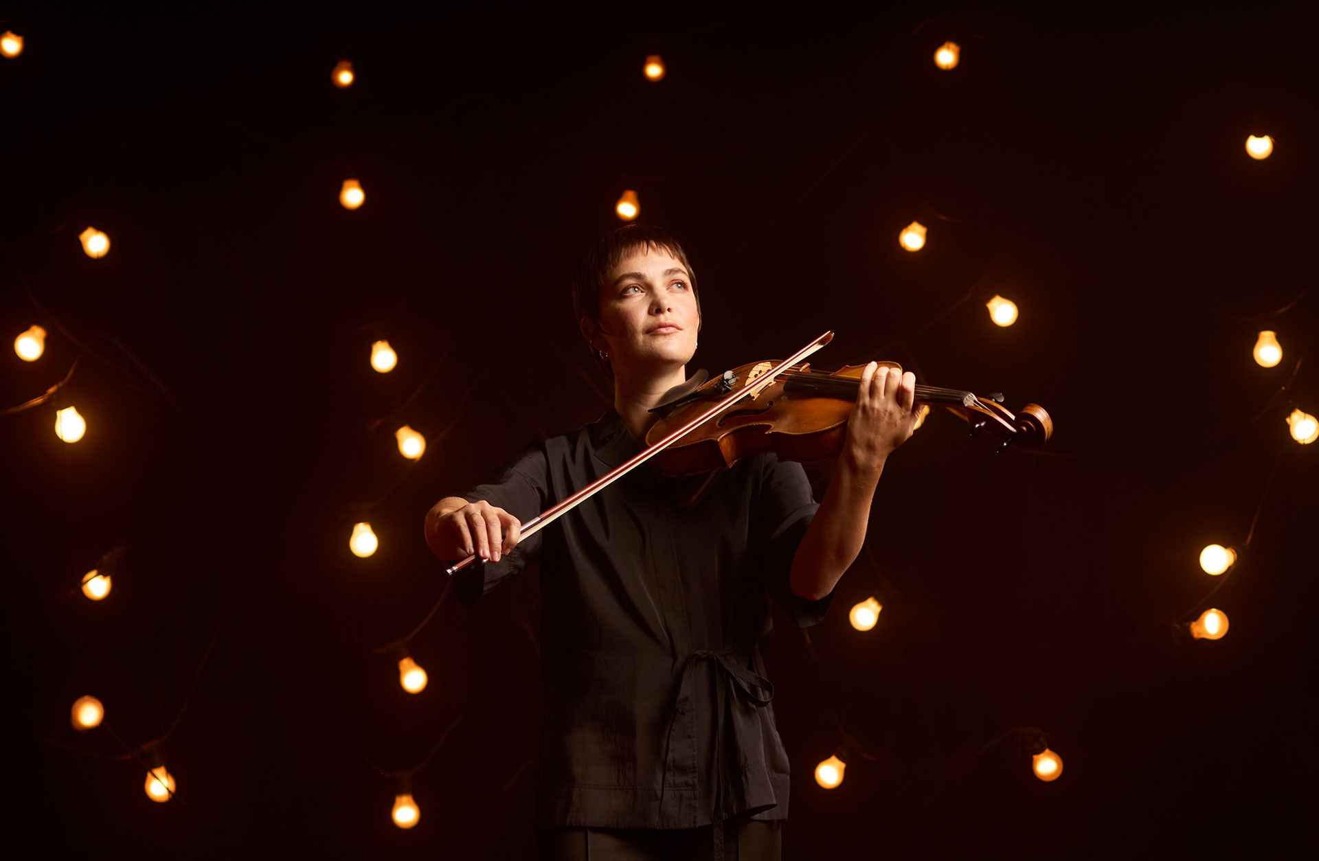 Kvinna med kort hår spelar viola mot en mörk bakgrund med lampor.