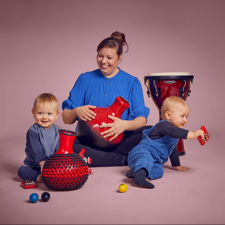 En kvinna sitter med två bebisar runt sig och de leker med instrument. De har blåa kläder mot ljusrosa bakgrund.