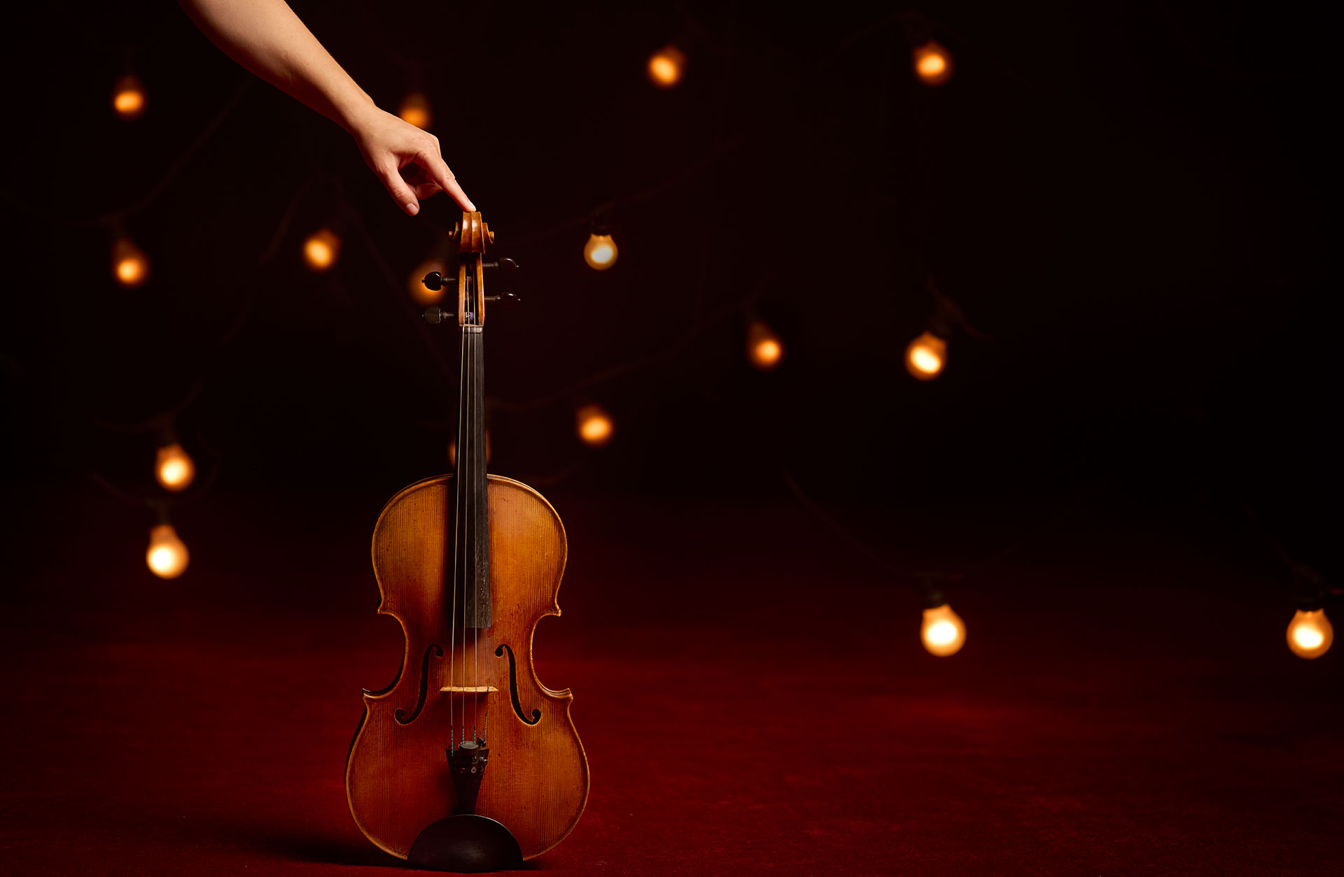Finger som håller i en violin som står på en röd matta med svart bakgrund med lampor.