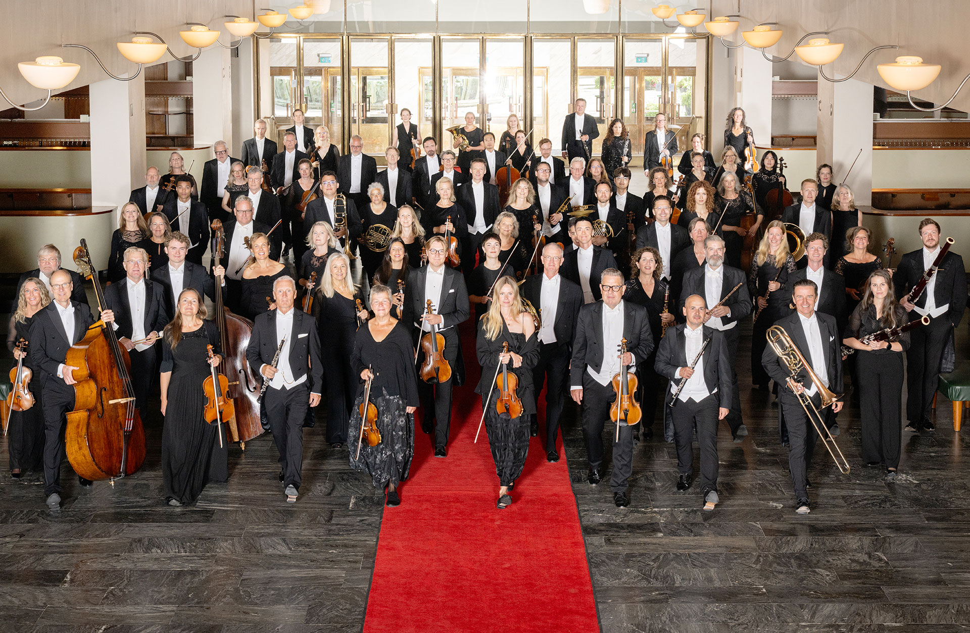 Musikerna i Göteborgs Symfoniker går mot kameran på röda mattan i konserthusets foajé.