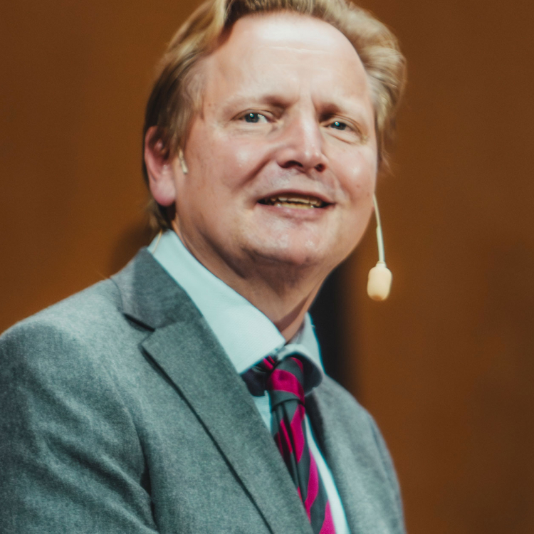 En blond man i grå kavaj som talar till en publik.