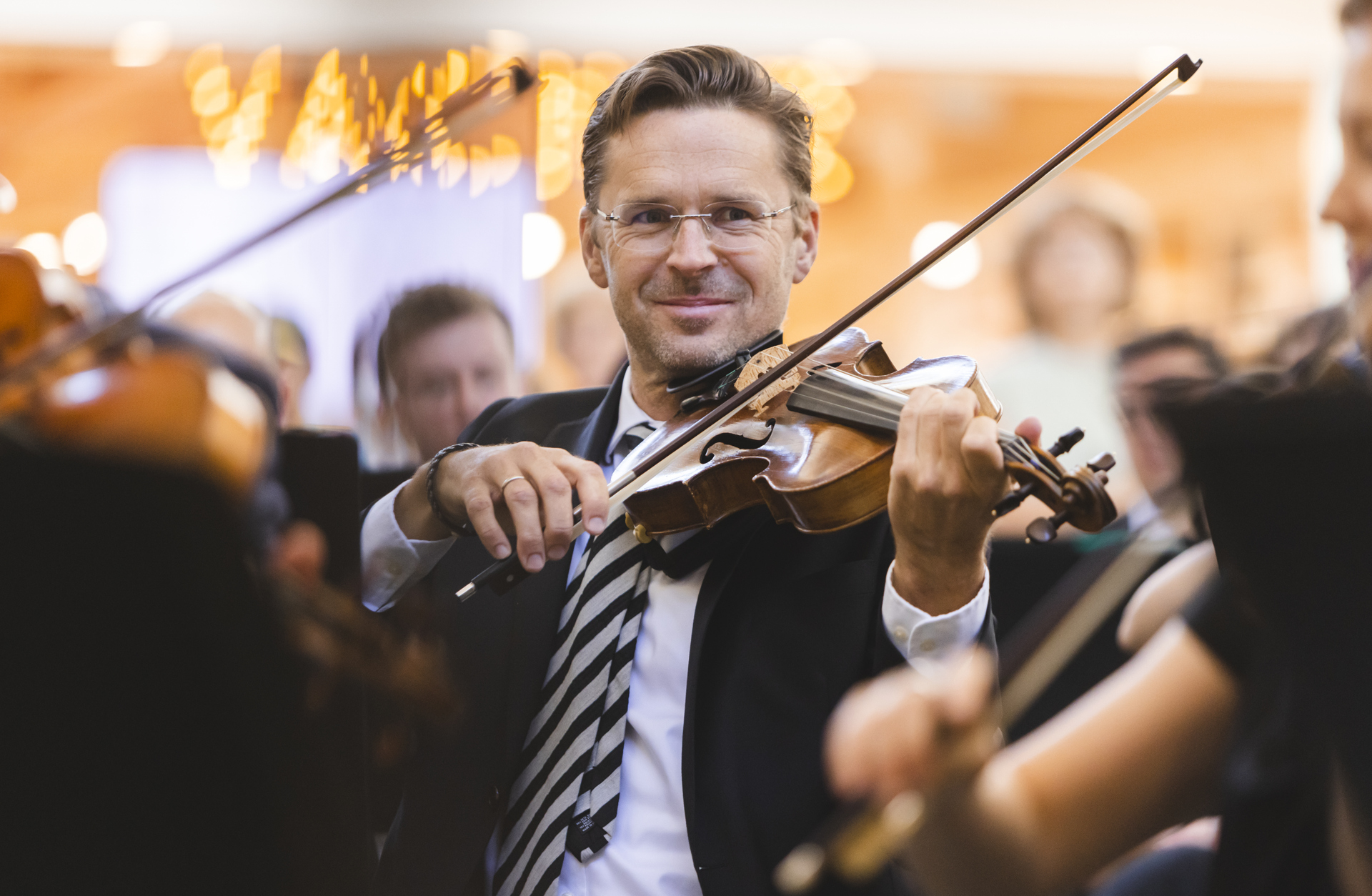 En glad man, klädd i svart kavaj och randig slips, tittar in i kameran och ler. Han spelar violin.