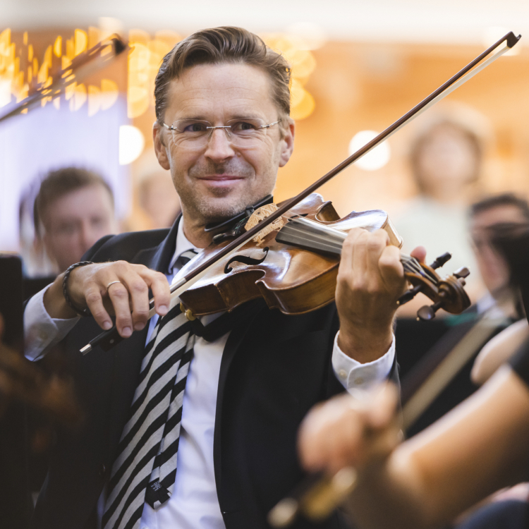 En glad man, klädd i svart kavaj och randig slips, tittar in i kameran och ler. Han spelar violin.