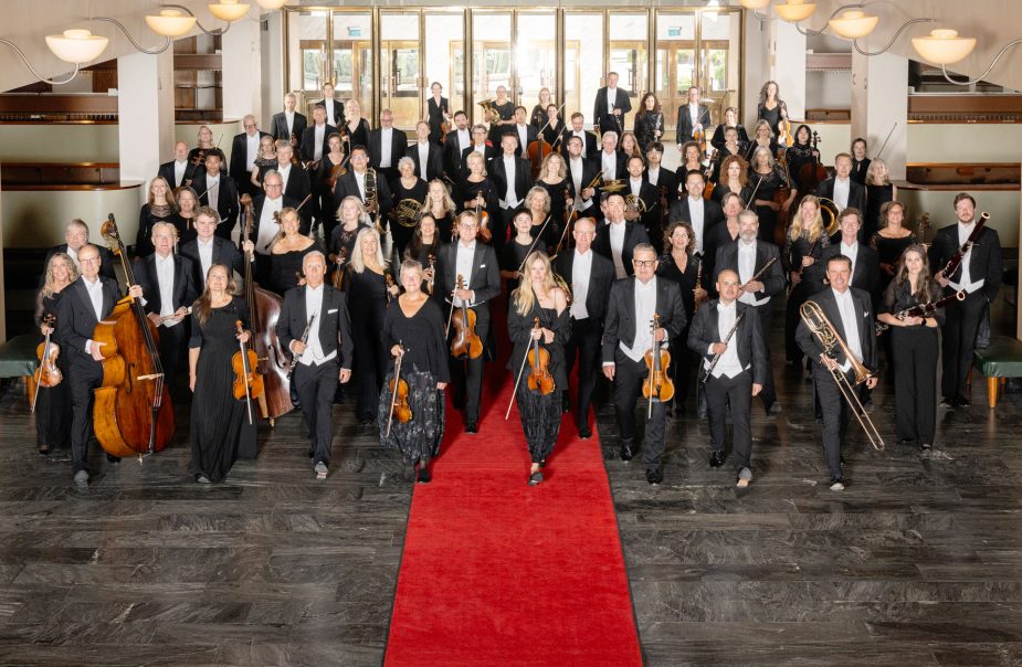 En konsertklädd symfoniorkester håller sina instrument och står i garderobshallen i Göteborgs Konserthus.