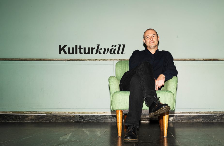 En svartklädd man sitter i en grön fåtölj med benen i kors. Han tittar in i kameran.